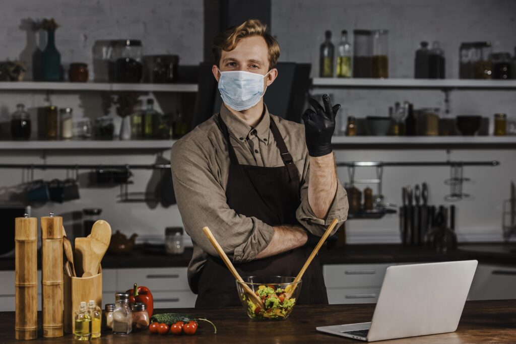 mid-shot-chef-with-mask-mixing-salad-ingredients-near-laptop